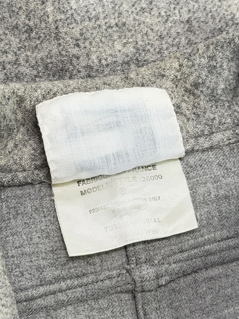 May include: Close-up of gray fabric with a white tag attached. The tag has text that includes "FABRIQUE EN FRANCE" and "MODEL/STYLE 26000". The fabric appears to be a woven material.