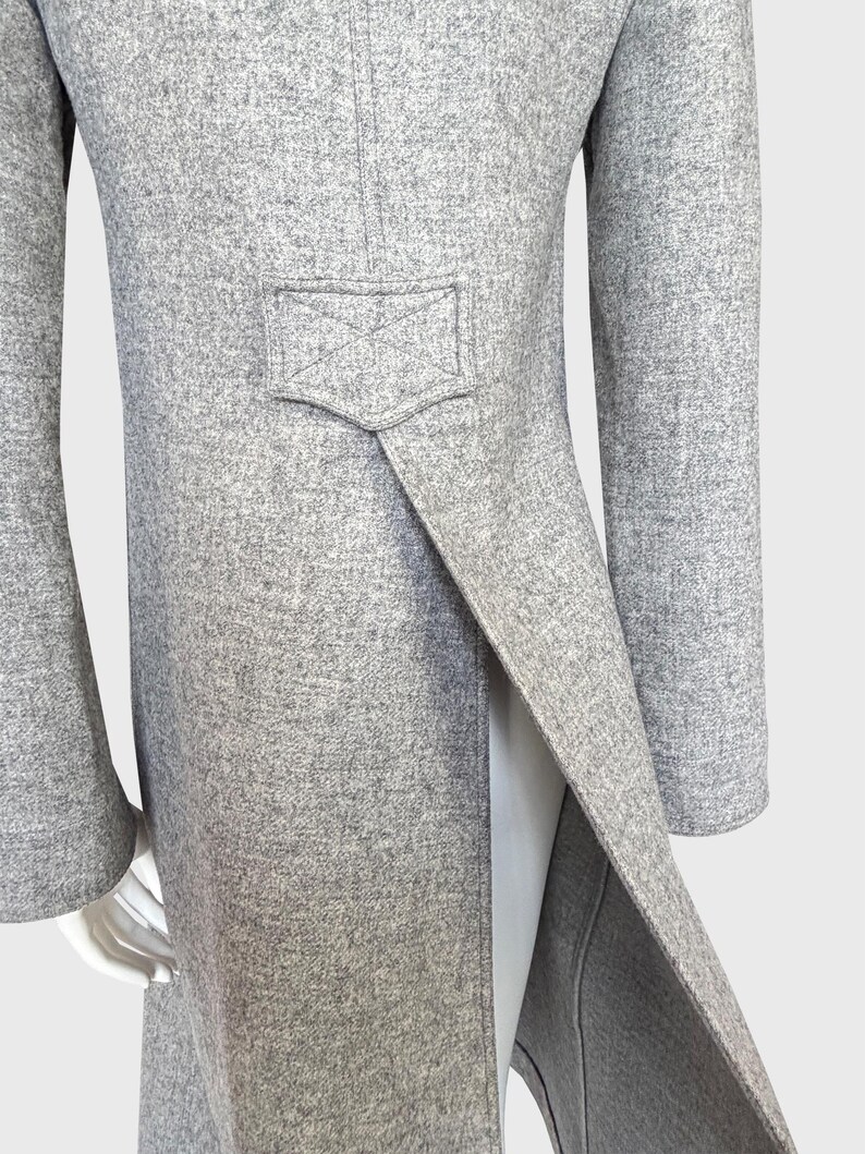 May include: A light gray wool coat with a split back and a small pocket detail. The coat has long sleeves and a tailored fit. The fabric has a subtle texture, and the coat appears to be well-constructed. The coat is displayed on a mannequin.