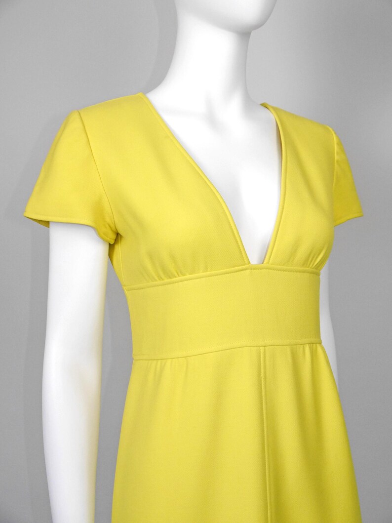 May include: A bright yellow dress with a deep V-neckline and short sleeves. The dress features a fitted waist and a flowing skirt. The fabric appears to be a smooth, solid color, and the dress is displayed on a mannequin.