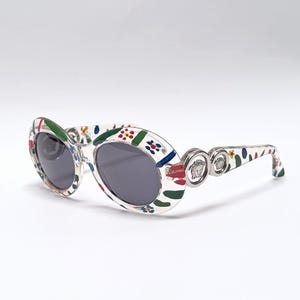 May include: Oval-shaped sunglasses with a white frame decorated with colorful floral and abstract patterns. The lenses are dark gray. The temples feature silver-toned Medusa head emblems. A stylish accessory for eye protection.