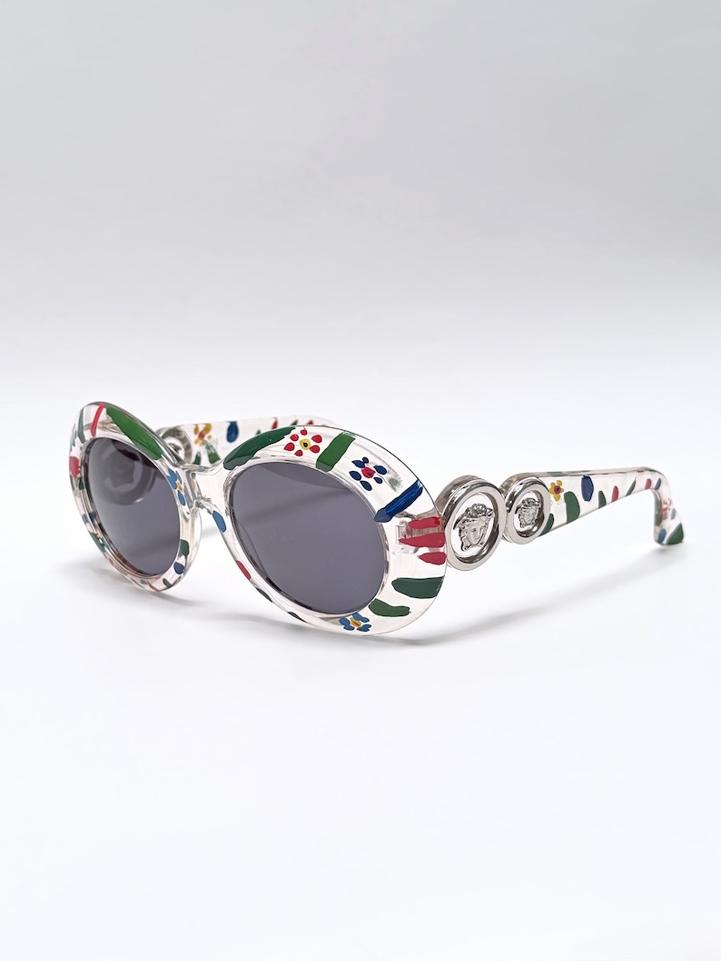 May include: Oval-shaped sunglasses with a white frame decorated with colorful floral and abstract patterns. The lenses are dark gray. The temples feature silver-toned Medusa head emblems. A stylish accessory for eye protection.