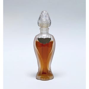 Op de afbeelding: Een heldere glazen parfumfles met een decoratieve stop. De fles heeft een unieke vorm, met een bredere basis die naar boven taps toeloopt. De vloeistof binnenin is amberkleurig. Een klein etiket is op de voorkant van de fles bevestigd.
