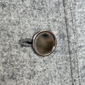 May include: Close-up of a gray wool coat with a large, shiny, silver button. The button is round with a smooth, reflective surface. The coat fabric has a textured appearance and a visible seam.