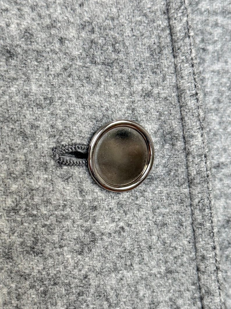 May include: Close-up of a gray wool coat with a large, shiny, silver button. The button is round with a smooth, reflective surface. The coat fabric has a textured appearance and a visible seam.