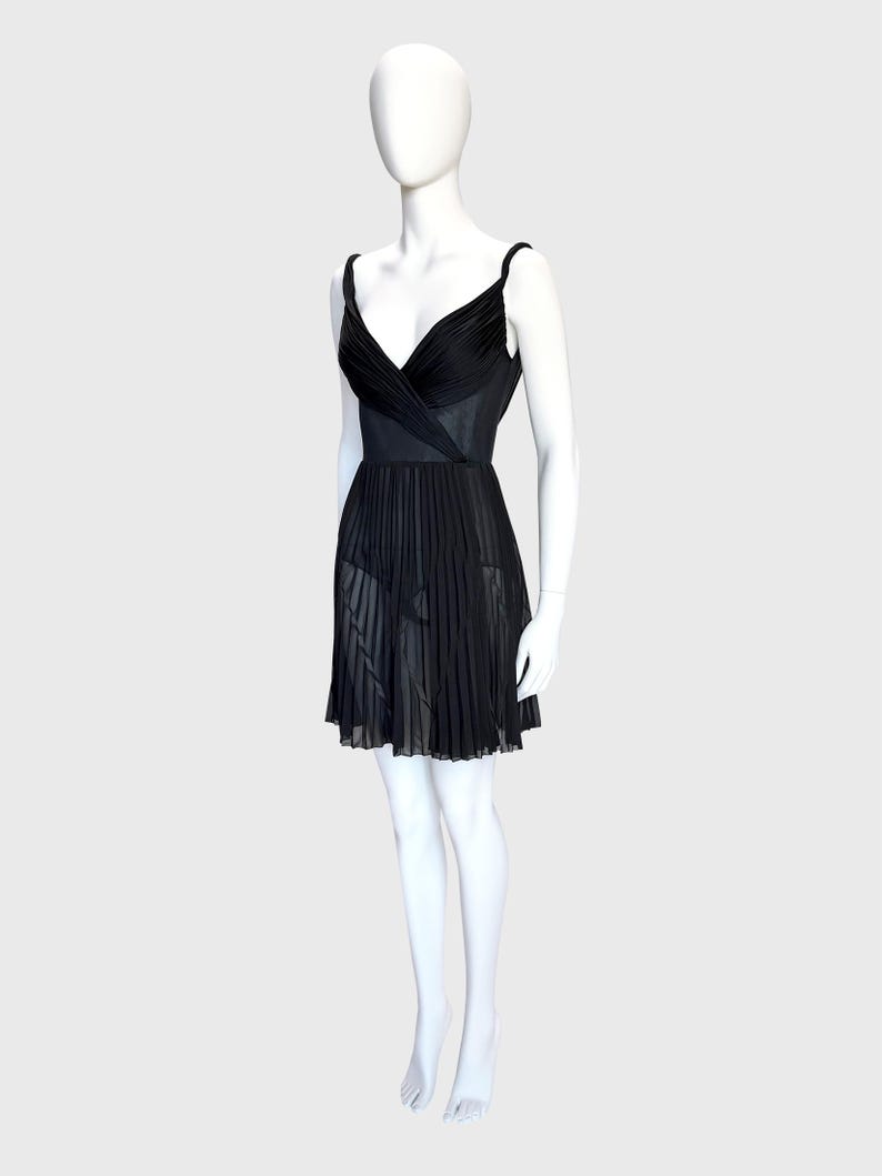 K&ouml;nnte beinhalten: Ein schwarzes Kleid auf einer Schaufensterpuppe. Das Kleid hat ein V-Ausschnitt-Oberteil mit Wickeldesign und d&uuml;nnen Tr&auml;gern. Der Rock ist plissiert und reicht bis zur Mitte des Oberschenkels. Das Kleid ist aus einem transparenten Stoff gefertigt.