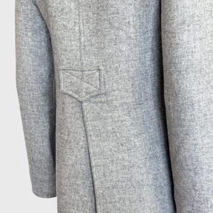 May include: Close-up of a light gray coat. The coat features a tailored design with a small, decorative pocket detail on the back. The fabric has a textured appearance, and the sleeves are long.