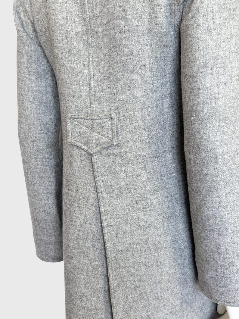 May include: Close-up of a light gray coat. The coat features a tailored design with a small, decorative pocket detail on the back. The fabric has a textured appearance, and the sleeves are long.