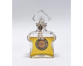 Può includere: Un flacone di profumo in vetro trasparente con un tappo a forma di cuore e un'etichetta che recita "Mitsouko Paris". Il flacone contiene un liquido giallo dorato. Un nappa decorativa pende dal collo del flacone.