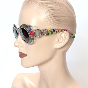 May include: Oval-shaped sunglasses with a clear frame and dark lenses. The frame is decorated with colorful floral and geometric patterns. Silver-toned Medusa head emblems are on the temples. The sunglasses are displayed on a mannequin head.
