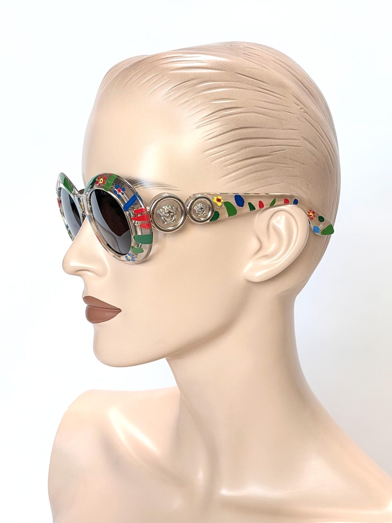 May include: Oval-shaped sunglasses with a clear frame and dark lenses. The frame is decorated with colorful floral and geometric patterns. Silver-toned Medusa head emblems are on the temples. The sunglasses are displayed on a mannequin head.
