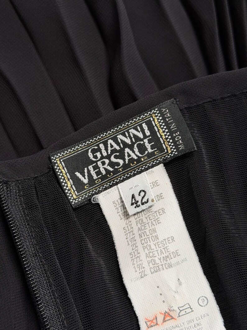 K&ouml;nnte beinhalten: Nahaufnahme eines schwarzen Kleidungsst&uuml;cks mit einem Etikett "GIANNI VERSACE COUTURE". Das Etikett zeigt Gr&ouml;&szlig;e 42 und Stoffzusammensetzung. Das Kleidungsst&uuml;ck hat eine plissierte Textur.