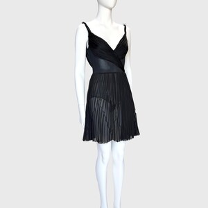 K&ouml;nnte beinhalten: Schwarzes Kleid mit plissiertem Rock und V-Ausschnitt. Das Kleid hat d&uuml;nne Tr&auml;ger und eine taillierte Taille. Der Rock ist halbtransparent, und das Kleid wird auf einer Schaufensterpuppe pr&auml;sentiert.