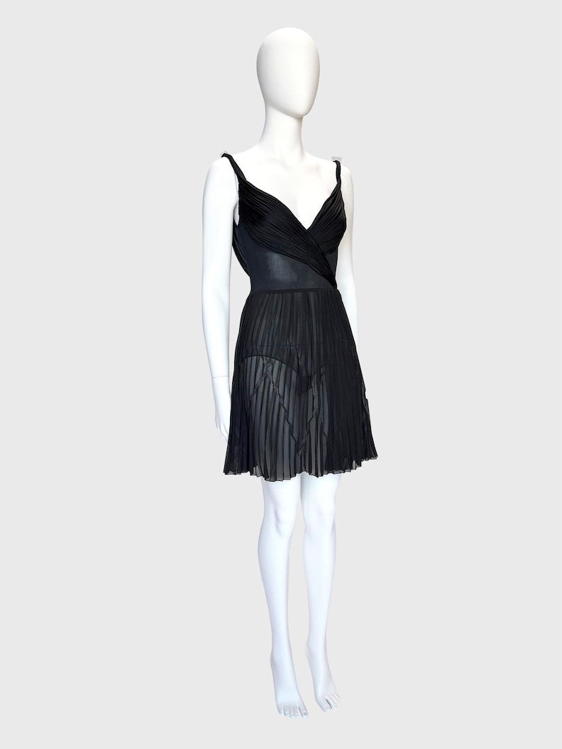 K&ouml;nnte beinhalten: Schwarzes Kleid mit plissiertem Rock und V-Ausschnitt. Das Kleid hat d&uuml;nne Tr&auml;ger und eine taillierte Taille. Der Rock ist halbtransparent, und das Kleid wird auf einer Schaufensterpuppe pr&auml;sentiert.