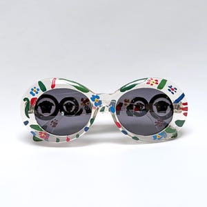 May include: Oval-shaped sunglasses with a clear frame, adorned with a colorful floral pattern in red, blue, and green. The lenses are dark, reflecting the surroundings. A stylish accessory for eye protection and fashion.
