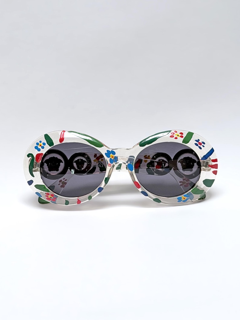 May include: Oval-shaped sunglasses with a clear frame, adorned with a colorful floral pattern in red, blue, and green. The lenses are dark, reflecting the surroundings. A stylish accessory for eye protection and fashion.