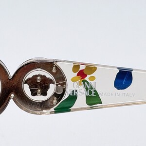 May include: Close-up of a pair of eyeglasses with a clear frame and decorative arms. The arms feature colorful abstract paint splatters and the text "GIANNI VERSACE MADE IN ITALY". The frame has a gold-colored Medusa head logo.