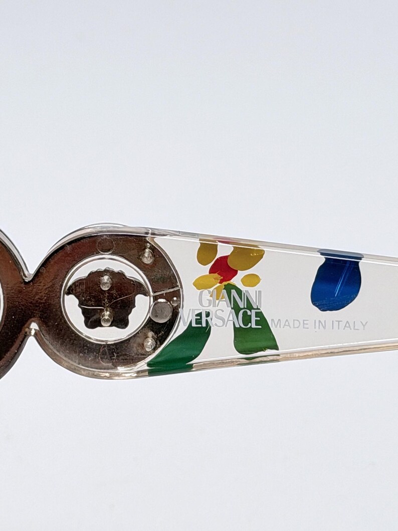 May include: Close-up of a pair of eyeglasses with a clear frame and decorative arms. The arms feature colorful abstract paint splatters and the text "GIANNI VERSACE MADE IN ITALY". The frame has a gold-colored Medusa head logo.