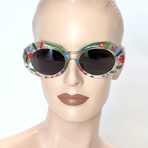 May include: Clear oval sunglasses with dark lenses. The frames are decorated with colorful floral designs in red, blue, and green. The sunglasses are displayed on a mannequin.