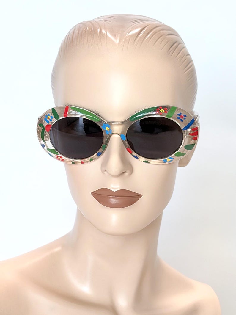 May include: Clear oval sunglasses with dark lenses. The frames are decorated with colorful floral designs in red, blue, and green. The sunglasses are displayed on a mannequin.