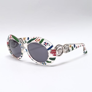 May include: Oval-shaped sunglasses with a white frame adorned with a colorful floral pattern. The lenses are dark gray, and the temples feature silver accents with a Medusa head logo. A stylish accessory.