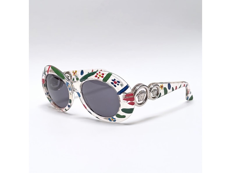 May include: Oval-shaped sunglasses with a white frame adorned with a colorful floral pattern. The lenses are dark gray, and the temples feature silver accents with a Medusa head logo. A stylish accessory.