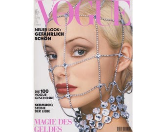 May include: A magazine cover featuring a woman wearing a silver chainmail face mask with rhinestones. The magazine is titled "Vogue" and the text "Neuer Look: Gefährlich Schön" is printed on the cover.