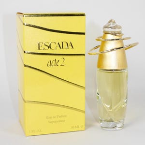 Puede incluir: Una caja amarilla con el texto "ESCADA acte 2" y un frasco de perfume de vidrio transparente con una tapa y anillos dorados. El frasco está etiquetado "Eau de Parfum Vaporisateur 30 ML e 1 FL. OZ."