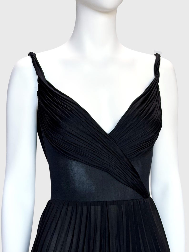 K&ouml;nnte beinhalten: Schwarzes Kleid mit V-Ausschnitt und plissiertem Rock. Das Oberteil hat ein drapiertes Design und das Kleid hat d&uuml;nne Tr&auml;ger. Das Kleid wird auf einer wei&szlig;en Schaufenquin vor hellem Hintergrund pr&auml;sentiert.