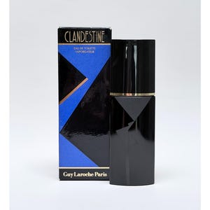 Può includere: Set di bottiglia e scatola di profumo neri. La scatola presenta la scritta "CLANDESTINE" in lettere dorate, con forme geometriche blu e nere. Il flacone di profumo è nero con una fascia dorata. Il testo "Guy Laroche Paris" è in basso sulla scatola.
