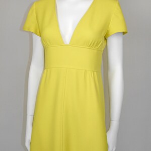 May include: A bright yellow dress with a V-neckline and short sleeves. The dress features a fitted waist and a flowing skirt. The dress is displayed on a white mannequin against a neutral background. The dress is made of a smooth, solid fabric.
