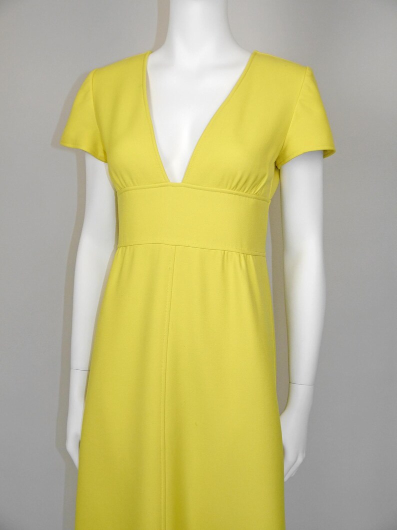 May include: A bright yellow dress with a V-neckline and short sleeves. The dress features a fitted waist and a flowing skirt. The dress is displayed on a white mannequin against a neutral background. The dress is made of a smooth, solid fabric.