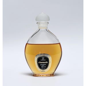 Könnte beinhalten: Ein runder Parfümflakon aus klarem Glas mit bernsteinfarbener Flüssigkeit. Der Flakon hat einen weißen, strukturierten Verschluss und ein schwarz-goldenes Etikett mit der Aufschrift eau de toilette chamade guerlain paris 100 ml 87% vol.
