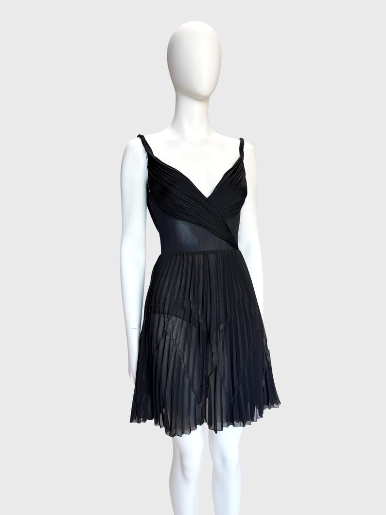 K&ouml;nnte beinhalten: Ein schwarzes Kleid mit V-Ausschnitt und Spaghettitr&auml;gern. Das Oberteil hat ein drapiertes Design und der Rock ist plissiert. Das Kleid wird auf einer wei&szlig;en Schaufensterpuppe vor wei&szlig;em Hintergrund pr&auml;sentiert.