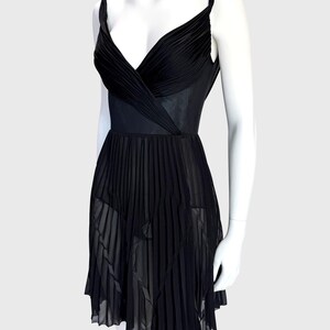 K&ouml;nnte beinhalten: Ein schwarzes Kleid mit einem plissierten Rock und einem V-Ausschnitt-Oberteil. Das Kleid hat d&uuml;nne Tr&auml;ger und eine taillierte Taille. Das Oberteil ist gerafft und drapiert, und der Rock hat ein flie&szlig;endes, plissiertes Design. Das Kleid wird auf einer Schaufensterpuppe pr&auml;sentiert.