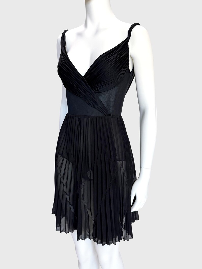 K&ouml;nnte beinhalten: Ein schwarzes Kleid mit einem plissierten Rock und einem V-Ausschnitt-Oberteil. Das Kleid hat d&uuml;nne Tr&auml;ger und eine taillierte Taille. Das Oberteil ist gerafft und drapiert, und der Rock hat ein flie&szlig;endes, plissiertes Design. Das Kleid wird auf einer Schaufensterpuppe pr&auml;sentiert.