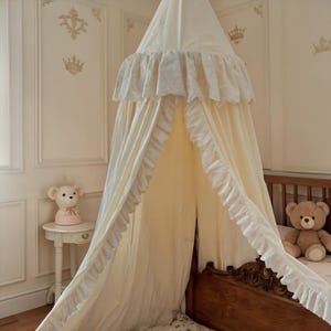 May include: A cream-colored fabric bed canopy featuring a gathered top section with a ruffled trim. The canopy drapes downward in long, flowing panels that are also finished with a ruffled edge, creating a soft, layered appearance over a bed.