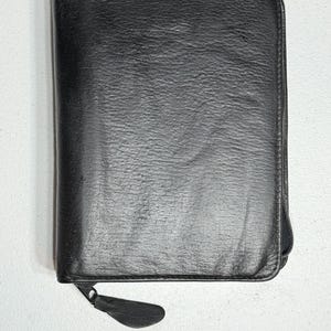 May include: A black leather-textured zippered portfolio case with a rectangular shape and rounded corners. The exterior features a visible grain pattern and a matching black zipper pull tab attached to the side closure mechanism.