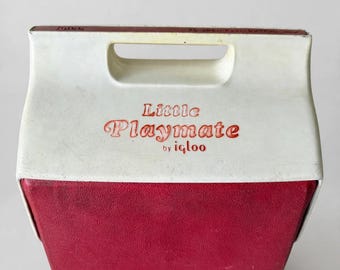 Puede incluir: Una nevera portátil Igloo Little Playmate de color blanco y rojo con un asa superior y el texto Little Playmate by igloo impreso en rojo en la parte frontal. La nevera tiene una forma rectangular cónica con una sección superior blanca y una base roja.