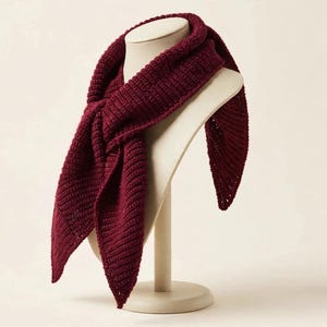 May include: A deep burgundy triangular scarf featuring a textured, ribbed crochet pattern. The scarf is draped in a knotted style, showcasing its pointed ends and consistent dark red yarn construction throughout the entire piece.