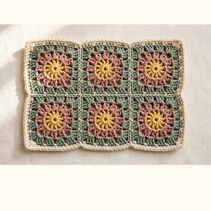 May include: A rectangular crochet piece composed of six joined granny squares. Each square features a central yellow flower, surrounded by dusty pink petals and sage green borders. The entire piece is edged with a cream-colored border.