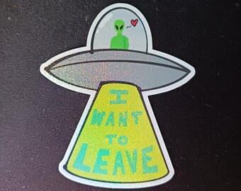 Può includere: Un adesivo raffigurante un UFO argentato con un alieno verde all'interno, un cuore sopra e la scritta "I WANT TO LEAVE" in verde e blu. L'adesivo ha un effetto olografico.