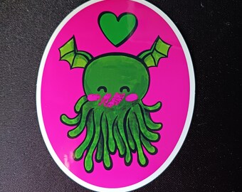 Può includere: Un adesivo con un mostro verde dei cartoni animati con tentacoli, ali e un cuore sopra la testa. Il mostro ha le guance rosa ed è ambientato su uno sfondo ovale rosa brillante con un bordo bianco. L'adesivo è probabilmente a scopo decorativo.