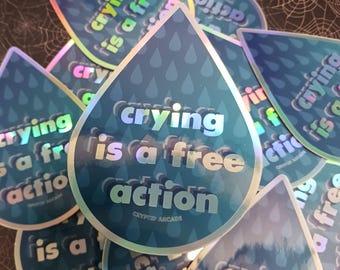 Può includere: Una collezione di adesivi olografici a forma di goccia. Gli adesivi sono blu con un motivo di piccole gocce di pioggia. Il testo "crying is a free action" è stampato sugli adesivi in un carattere riflettente e iridescente. È stampato anche il testo "Crypto Arcade".