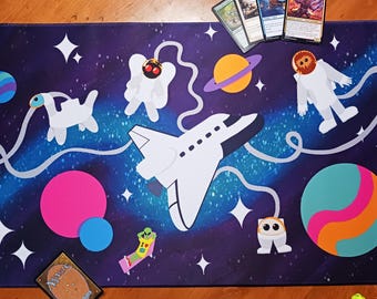 Può includere: Un tappetino da gioco rettangolare con uno sfondo spaziale blu scuro, uno space shuttle bianco, vari pianeti in rosa, arancione e turchese, e personaggi dei cartoni animati in tute spaziali bianche. Il design include stelle e una linea bianca.