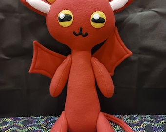 Può includere: Un giocattolo di peluche rosso con corna, ali e coda. Il giocattolo ha grandi occhi rotondi e una piccola bocca sorridente. È seduto su una superficie con un motivo colorato.