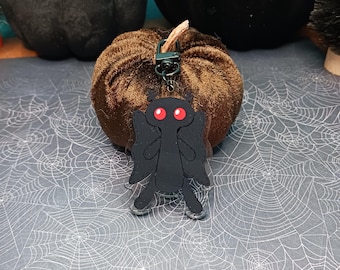 May include: Black and red acrylic charm of a creature with wings, hanging from a brown velvet pumpkin. The charm is attached to a black metal key chain.