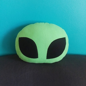 May include: Green alien-shaped cushion with black eyes. The cushion is made of soft fabric and is perfect for cuddling.