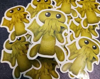 Può includere: Una collezione di adesivi raffiguranti una creatura a fumetti verde oliva con grandi occhi, tratti del viso simili a tentacoli e piccole ali. Gli adesivi sono fustellati con un bordo bianco e sono sparsi su una superficie scura.