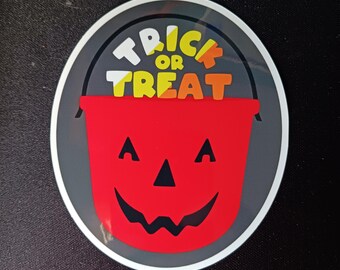 Può includere: Un adesivo circolare con un secchio di zucca rosso con una faccia sorridente. Le parole "TRICK OR TREAT" sono sopra il secchio in lettere bianche, gialle e arancioni. L'adesivo ha un bordo grigio e si trova su uno sfondo nero.