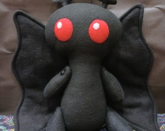 Può includere: Giocattolo di peluche nero con grandi occhi rossi e ali di farfalla. Il giocattolo è seduto su una superficie con un motivo colorato.
